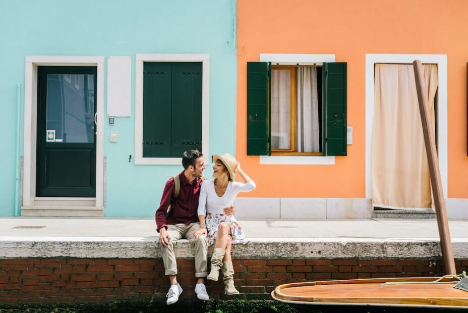 Am Hafen sitzendes, lachendes Paar verbringt günstige Flitterwochen in Europa. 